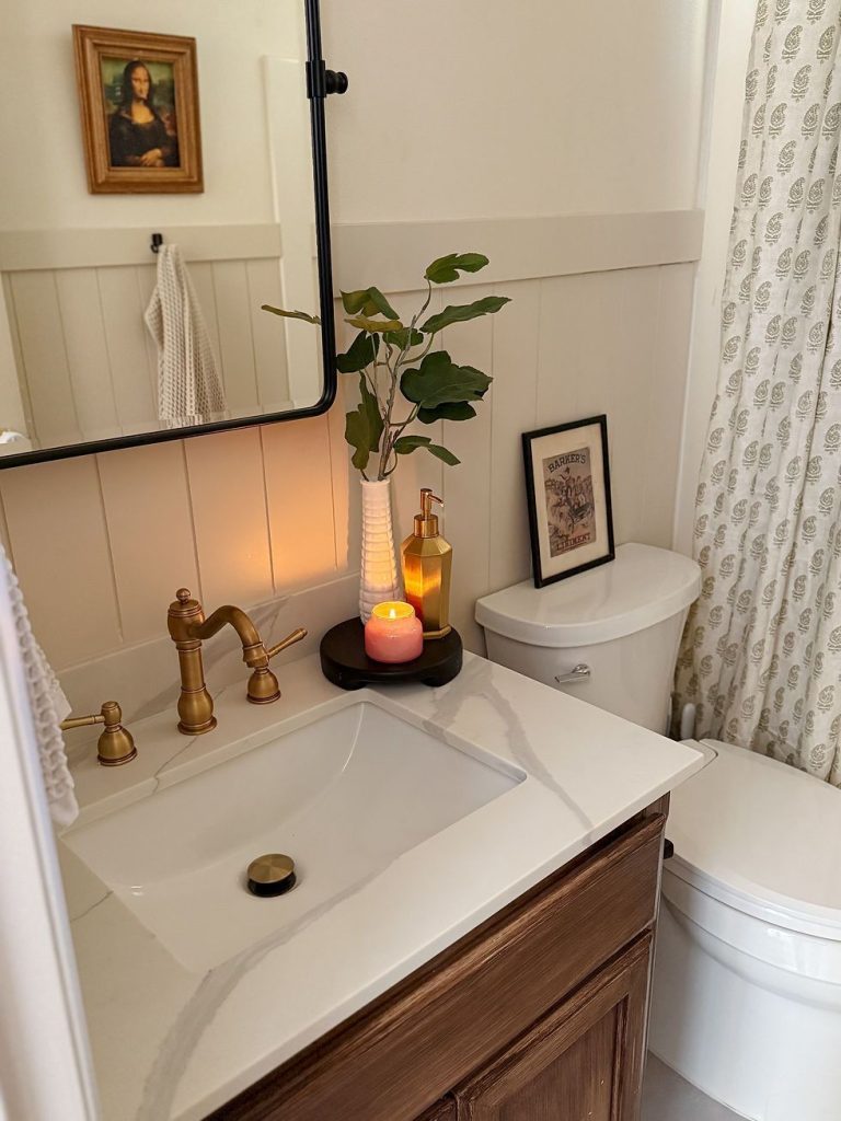 Warm and cozy small guest bathroom with beadboard wainscoting, a brass bridge faucet, white marble countertop, a lit pink candle on a wooden tray, a gold soap dispenser, a tall white vase with green stems, and a block print shower curtain.