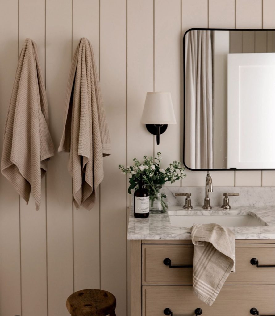 Serene neutral guest bathroom with warm beige shiplap paneling, a sand-toned painted vanity with black knobs, a black-framed rectangular mirror, a single black wall sconce, white marble countertop, ribbed linen hand towels hanging on the wall, and a small bunch of white flowers in a glass jar.