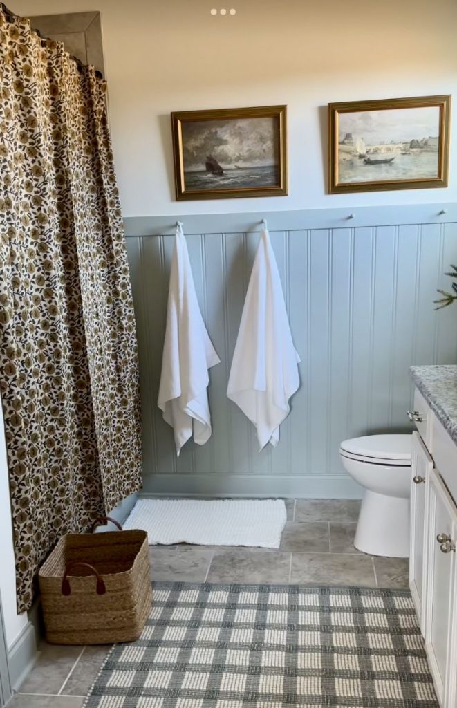 Refreshed guest bathroom with sage blue painted beadboard wainscoting, two white towels hanging on wall hooks, a floral block print shower curtain, gold-framed vintage landscape art above the wainscoting, a woven tote basket on the floor, and a plaid runner rug.