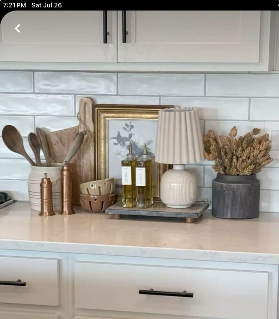 White kitchen counter styled with a grouped arrangement including a striped ceramic utensil crock with wooden spoons, a wooden cutting board, copper salt and pepper grinders, a colander-style berry bowl, two glass oil and vinegar dispensers on a small footed tray, a small ribbed ceramic table lamp with a cream pleated shade, and a dark stone pot with dried wheat stems, against a white subway tile backsplash.