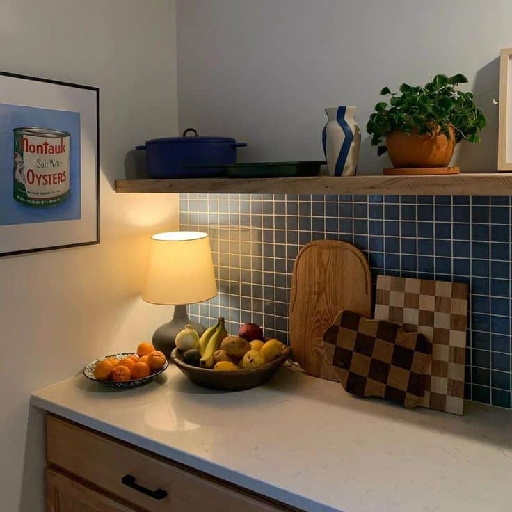 Eclectic kitchen counter with navy blue mosaic tile backsplash, a wooden floating shelf above holding a blue Le Creuset pot, a striped ceramic vase, and a potted green plant in a terracotta pot, with the counter below holding a small table lamp, a wooden fruit bowl with bananas, pears, and apples, a speckled ceramic plate with mandarin oranges, and two cutting boards including a checkered wood design, beside a black-handled cabinet drawer.