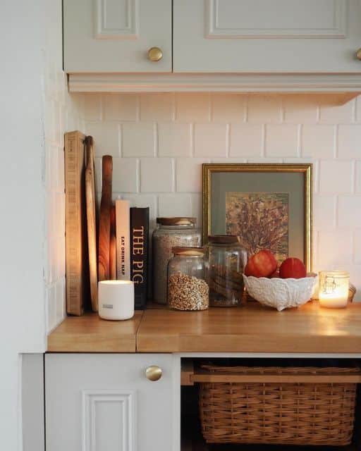 Warm rustic kitchen counter with cream Shaker cabinets and brass knob hardware, styled with several wooden and olive cutting boards propped upright, two large glass storage jars filled with grains, a white ceramic bowl with red apples, a small gold-framed vintage botanical print, a glowing white candle, and a small glass votive candle, on a butcher block countertop, with a wicker basket below.