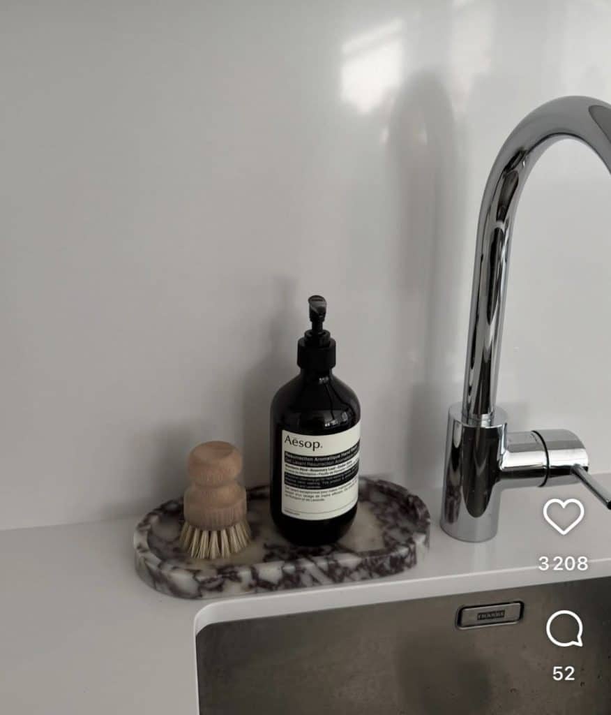 Minimalist modern kitchen sink area with a chrome gooseneck faucet, styled with a small round purple-veined marble tray holding a dark amber glass soap pump dispenser and a small natural wooden dish brush with bristle head, on a plain white countertop with a clean grey backsplash.