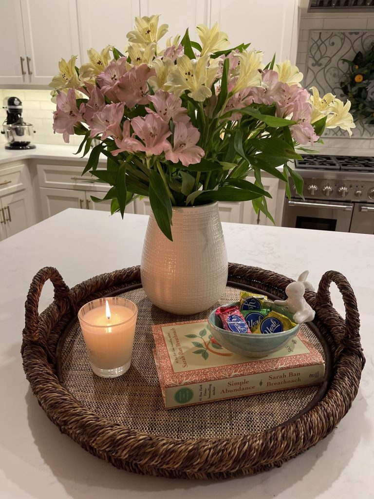 White kitchen island styled with a large round dark woven rattan tray holding a white ribbed ceramic vase with a large bunch of pink and yellow alstroemeria flowers, a lit glass candle, two stacked vintage hardcover books, and a small decorative bowl with wrapped candies and a ceramic bunny figurine, in a white kitchen with subway tile backsplash and stainless steel range.