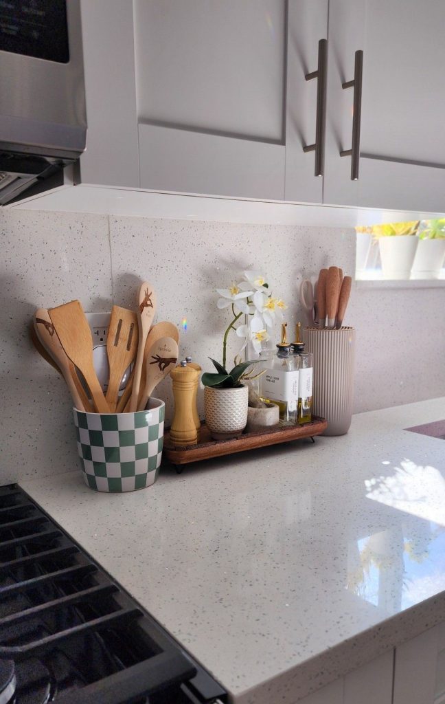 Modern white kitchen counter beside a stove styled with a green and white checkerboard ceramic utensil crock filled with wooden spatulas, and a narrow dark wooden footed tray holding a yellow wooden pepper grinder, a small textured ceramic succulent pot, two glass oil and vinegar bottles with gold dispenser tops, and a ribbed cream knife block, with a white quartz speckled countertop and matching backsplash.