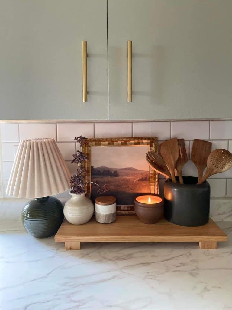 Sage green kitchen with brass hardware and white subway tile backsplash, with a counter styled with a large rectangular blonde wooden riser holding a dark green ribbed ceramic table lamp with a cream pleated shade, a small white and cream ceramic vase with dried purple stems, a gold-framed vintage landscape oil painting, a white and grey lidded ceramic jar, a glowing amber candle in a clay pot, and a dark matte utensil crock with wooden spatulas and spoons.