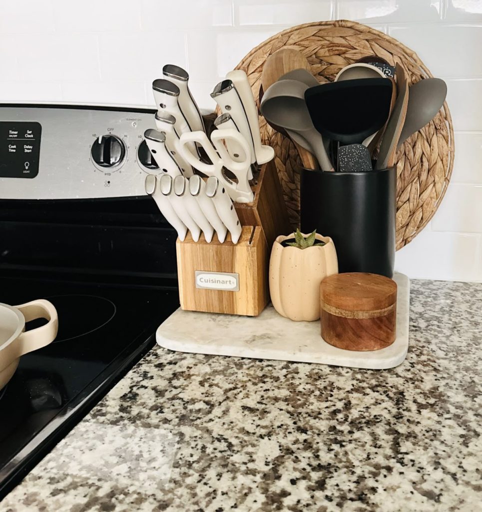 Modern kitchen counter near a stove styled with a marble tray holding a wooden Cuisinart knife block with cream-handled knives, a matte black utensil holder, a small pumpkin-shaped ceramic pot with a succulent, and a round wooden jar with lid, beside a woven rattan placemat leaning against the backsplash.