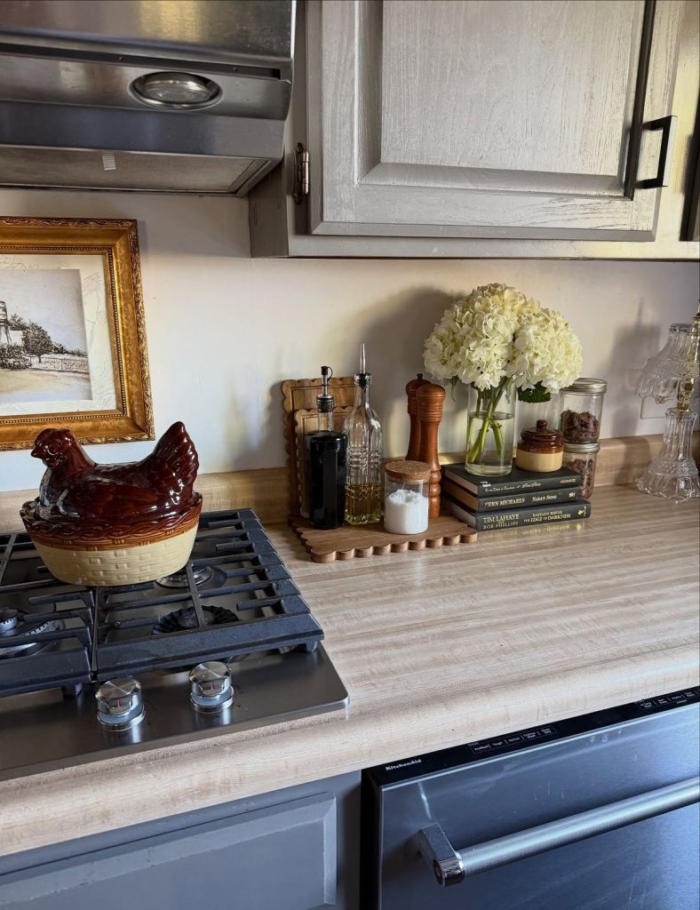 Cottagecore-inspired kitchen counter with grey-white painted cabinets and a wood-look benchtop, styled with a vintage ceramic hen pot on the stovetop, a gold-framed sepia landscape print leaning against the backsplash, two glass olive oil and vinegar bottles, a tall wooden pepper grinder, a glass jar with a cork lid, a clear glass vase with white hydrangeas, and a small decorative wooden scalloped tray holding kitchen items, with a vintage-style gold-framed art print to the left.