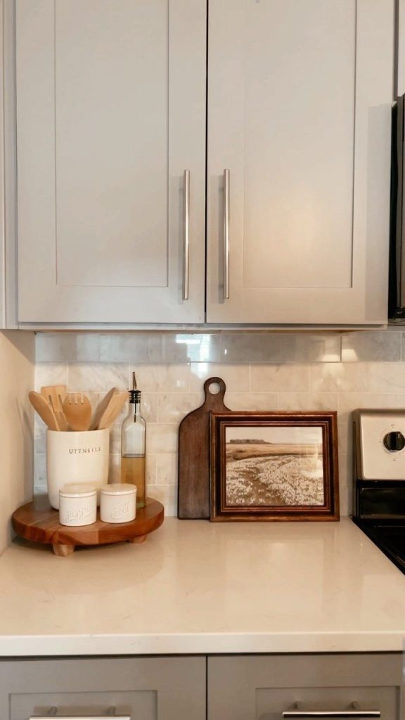 Soft white kitchen counter with a marble-look backsplash, styled with a round wooden lazy Susan holding a white ceramic utensil crock labeled utensils with wooden spatulas, a glass oil bottle with a pour spout, and two small white ceramic salt and pepper pots, beside a dark walnut cutting board and a small vintage landscape print in a dark frame leaning against the backsplash.