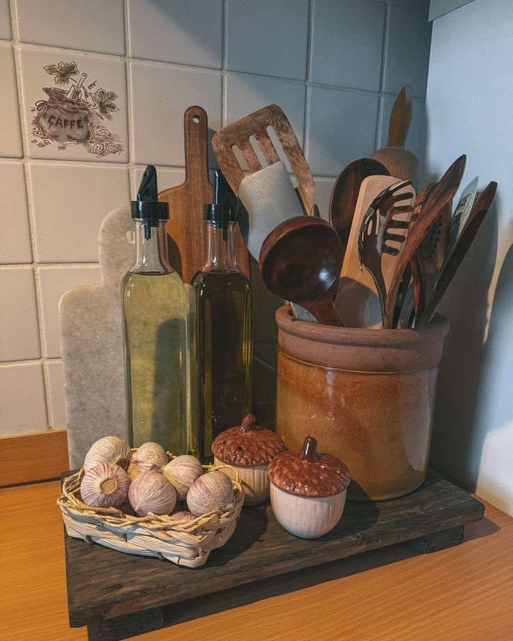 Rustic farmhouse kitchen counter styled on a dark weathered wooden riser holding two tall glass olive oil and vinegar bottles, a large terracotta-glazed ceramic utensil pot filled with an abundance of wooden spoons, spatulas, and other cooking tools, a small wicker basket with fresh garlic heads, two ceramic acorn salt and pepper shakers, and a small marble cutting board propped upright, beside vintage white square kitchen tiles with a hand-painted coffee motif.