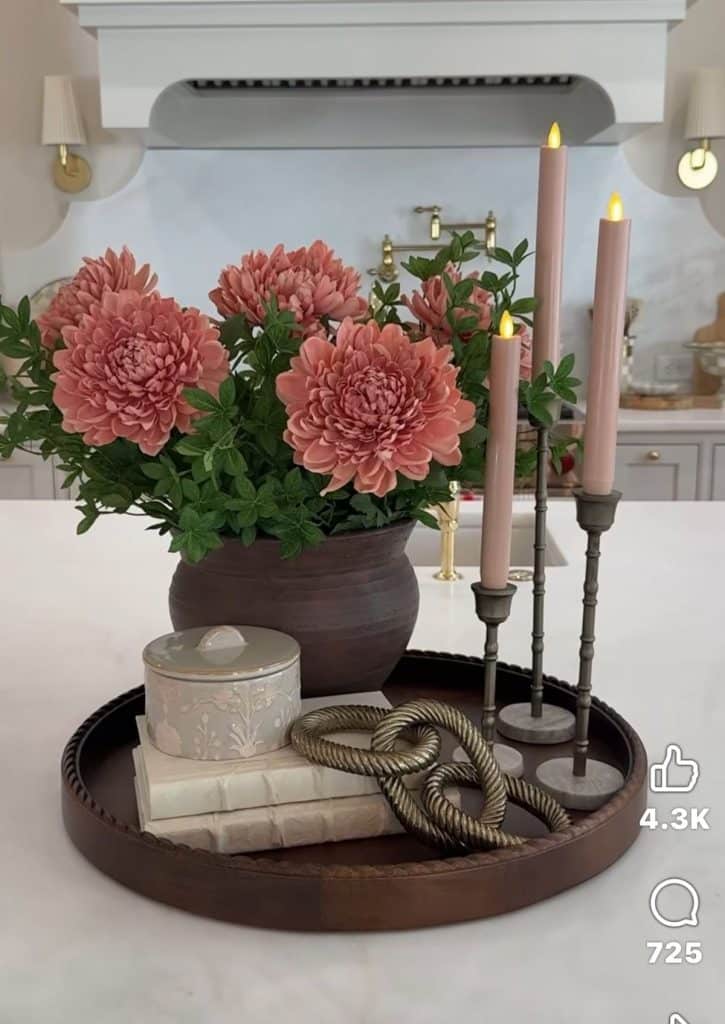 Elegant kitchen island counter styled with a large dark round metal tray holding a large dark clay bowl filled with full blooms of pink chrysanthemum or peony flowers, three varying-height antique bronze taper candle holders with pale blush pink flameless taper candles, a small floral embossed ceramic lidded box, two stacked linen-covered books, and a decorative twisted gold rope ornament, in a white kitchen with a decorative range hood and brass sconce lighting.
