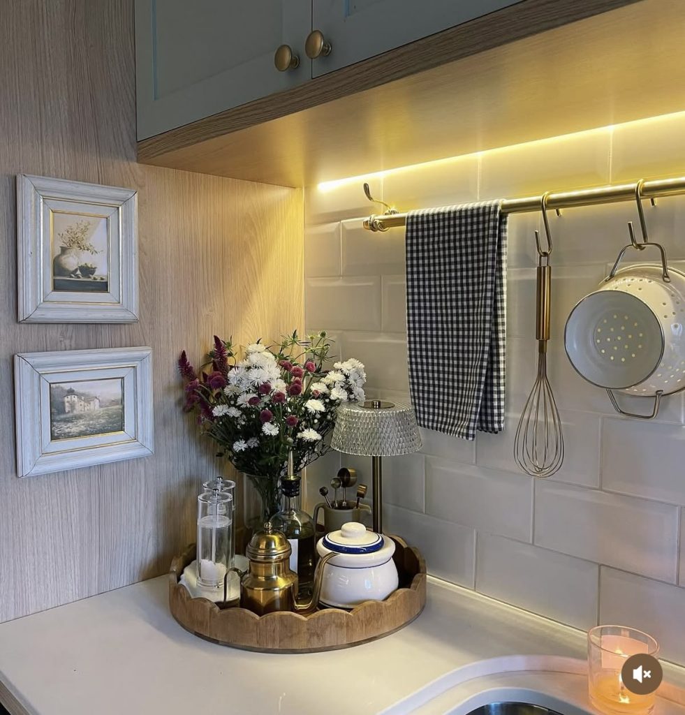 French country-inspired kitchen counter corner with sage blue-green upper cabinets with brass knob hardware, warm wood paneled backsplash walls with small framed vintage still-life art prints, warm under-cabinet LED strip lighting, and a brass S-hook rail holding a black gingham tea towel, a whisk, and a small colander, with the counter below holding a round scalloped-edge wooden tray styled with a small crystal table lamp, a bunch of mixed wildflowers in a glass vase, a brass French press coffee maker, a glass sugar jar, a small white ceramic sugar pot with blue trim, a candle, and a small lit votive glass.