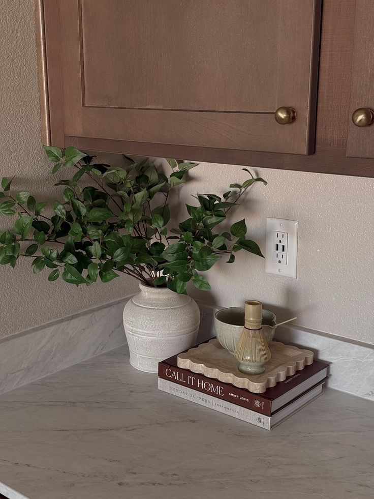 Warm kitchen counter corner with timber cabinets and brass knobs, styled with a large textured white ceramic vase holding leafy green stems, a stack of hardcover coffee table books including Call It Home, a scalloped stone tray with a ceramic matcha bowl and whisk, on a white marble-look countertop.