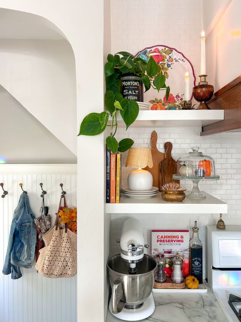 Eclectic and maximalist kitchen with open white floating shelves above a marble countertop, styled with a trailing pothos plant, vintage salt tin, floral decorative plate, amber glass candlestick, a small table lamp with rattan shade, stacked plates, wooden and olive cutting boards, a glass cloche with seasonal decor, a KitchenAid stand mixer, and a canning handbook on the counter below.