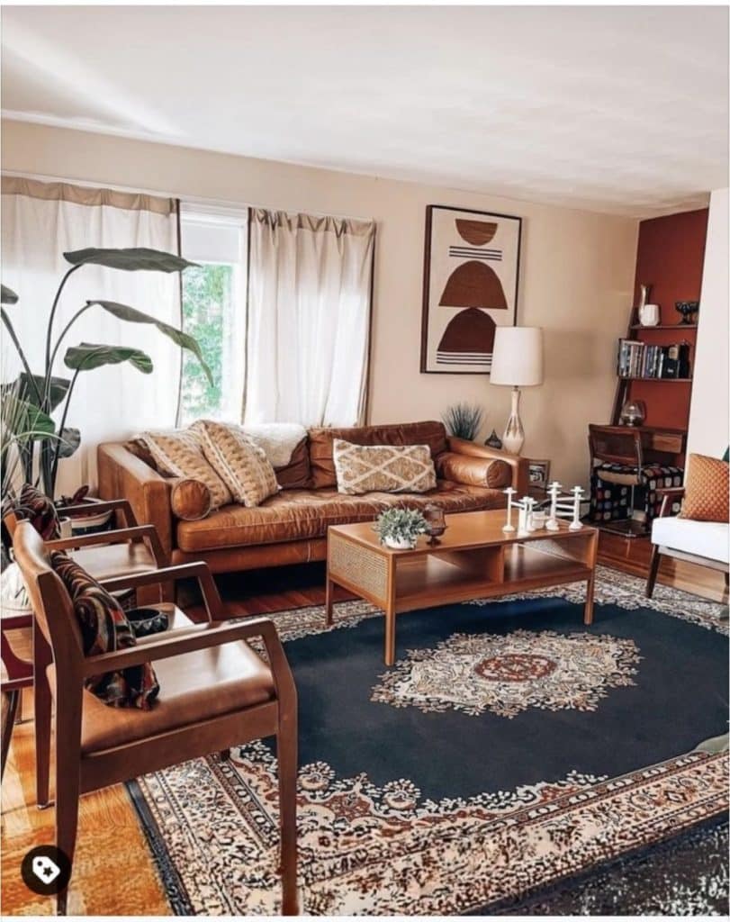 Mid century modern living room with a tan leather sofa, two wooden frame armchairs with leather seats, a cane coffee table, a large navy Persian rug, abstract geometric art print above the sofa, a banana leaf plant, and a terracotta accent wall with bookshelves