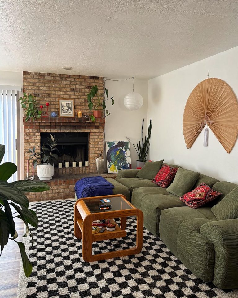 Mid century modern living room with a large olive green corduroy sectional sofa, a rounded wood and glass coffee table, a black and white checkerboard shag rug, a brick fireplace with plants and candles on the mantle, a large dried palm leaf wall hanging, houseplants throughout, and a paper globe pendant light