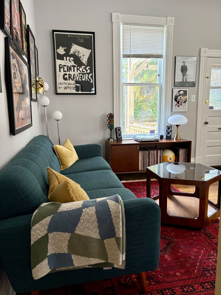 Mid century modern living room with a teal fabric sofa, yellow fringe throw pillows, a wooden credenza holding a record player and vinyl collection, a globe floor lamp, a red Persian rug, and a gallery wall of black and white photography and art prints