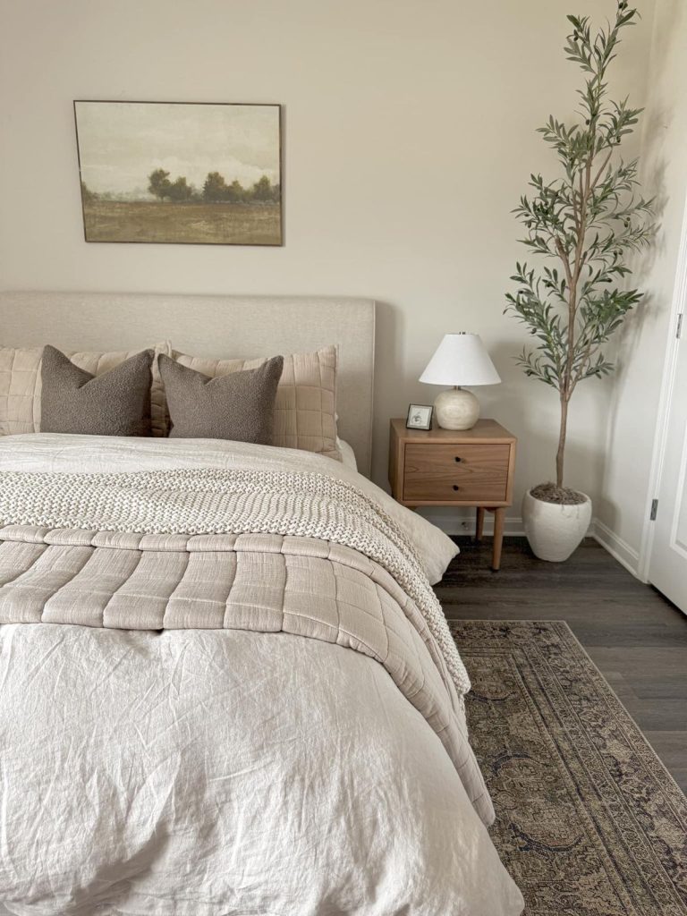 Minimalist bedroom with neutral linen bedding, olive tree in white pot, wood nightstand with ceramic lamp, and vintage rug on dark hardwood floors