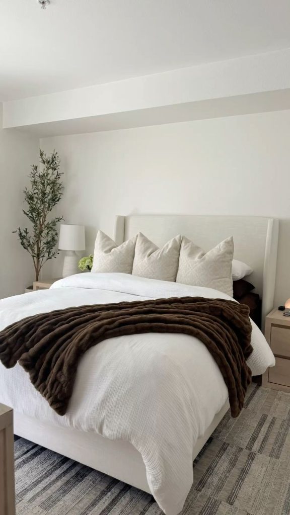 Clean minimalist bedroom with white upholstered wingback headboard, cream patterned euro pillows, rich brown faux fur throw, faux olive tree, and neutral area rug