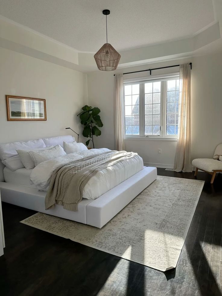 Bright minimalist bedroom with low platform bed, white bedding, woven pendant light, fiddle leaf fig, beige vintage style rug on dark hardwood floors, and neutral linen curtains