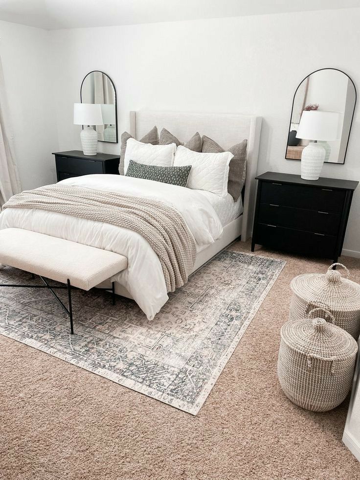 Neutral minimalist bedroom with cream upholstered headboard, white and tan bedding, black nightstands, arched mirrors, boucle bench, and woven storage baskets