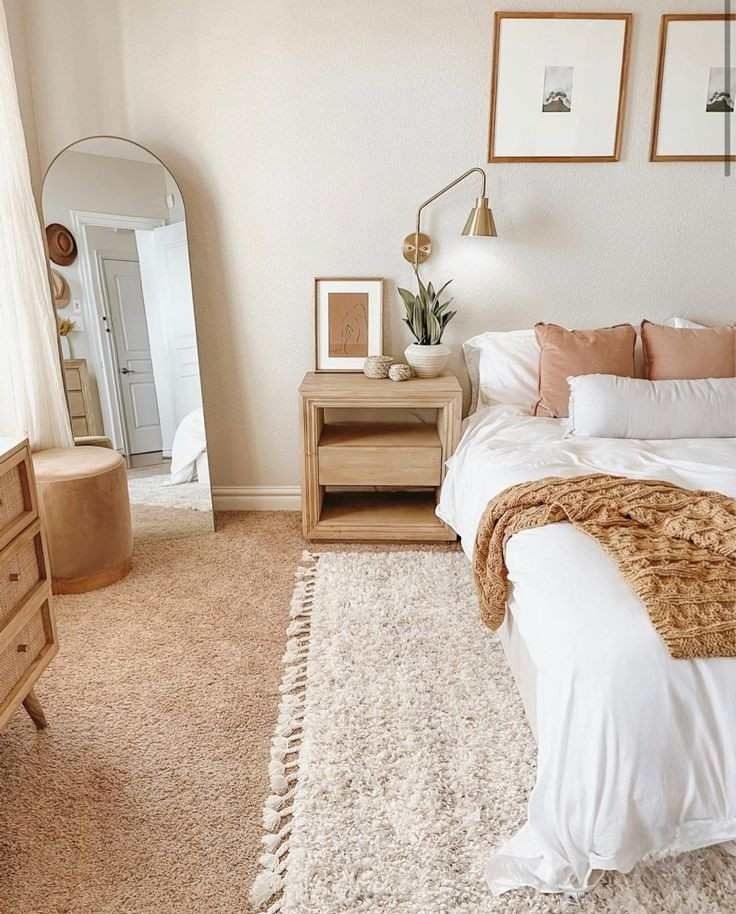 Warm minimalist bedroom with arched gold frame floor mirror, light wood open nightstand, brass wall sconce, white and blush bedding with ochre knit throw, and framed mountain prints
