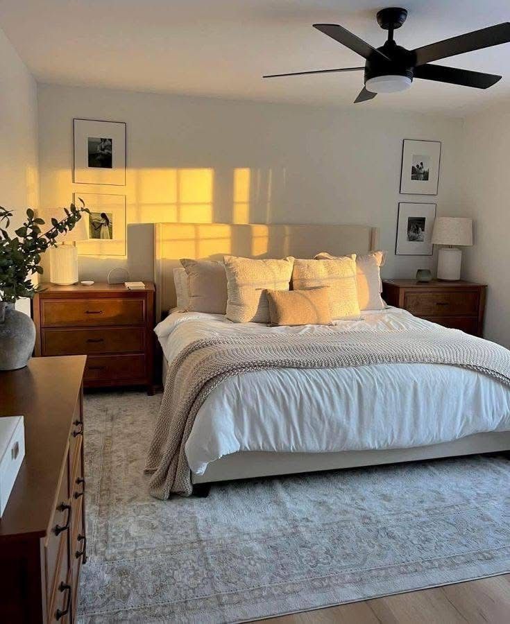 Golden hour minimalist bedroom with beige upholstered headboard, warm yellow accent pillows, walnut nightstands, black ceiling fan, framed black and white photos, and vintage rug