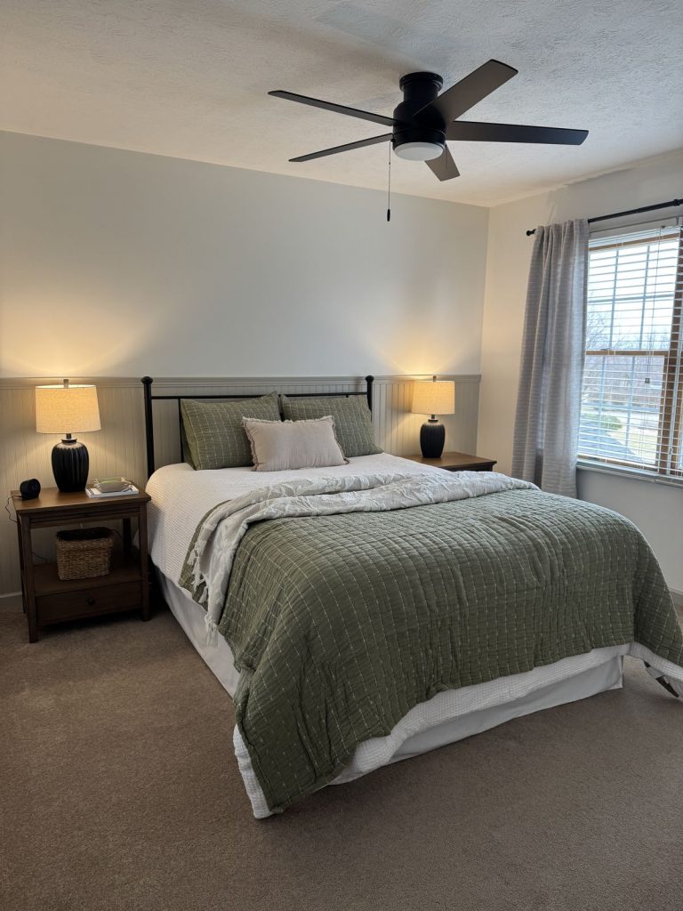 Minimalist bedroom with black iron bed frame, sage green quilt and matching pillows, beadboard wainscoting, matching wooden nightstands with dark lamps, and ceiling fan