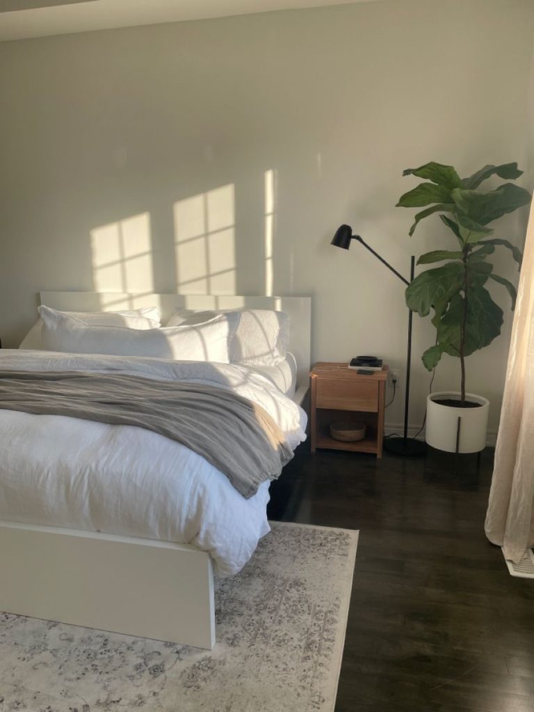 Bright minimalist bedroom with white platform bed, linen bedding, warm grey stripe throw, wooden open nightstand with reading lamp, tall fiddle leaf fig plant, and vintage area rug