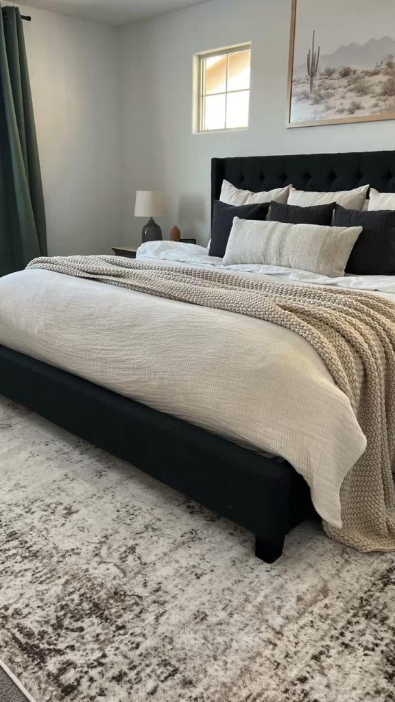 Minimalist bedroom with black tufted headboard, white and neutral bedding layered with a chunky knit throw, dark curtains, and vintage style area rug