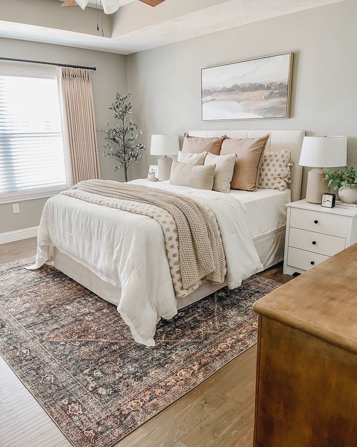 eutral minimalist bedroom with cream upholstered headboard, warm toned pillows, faux olive tree, white nightstands, and a traditional patterned area rug