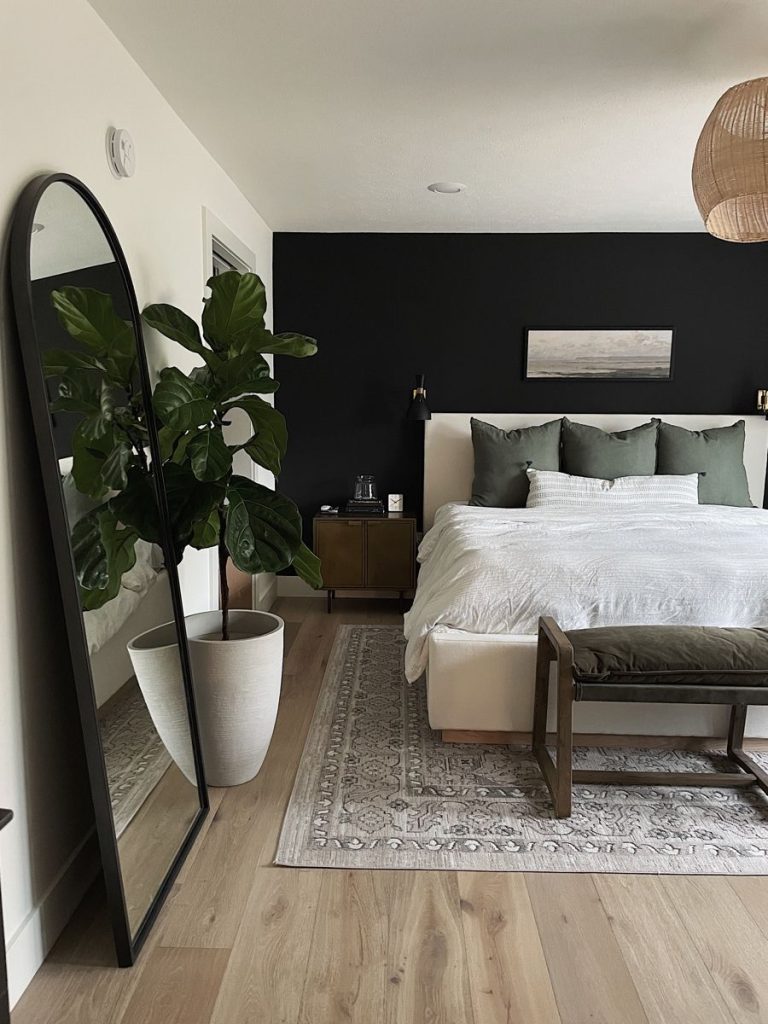 Modern minimalist bedroom with black accent wall, white upholstered bed, sage green pillows, arched floor mirror, fiddle leaf fig plant, and bench at foot of bed