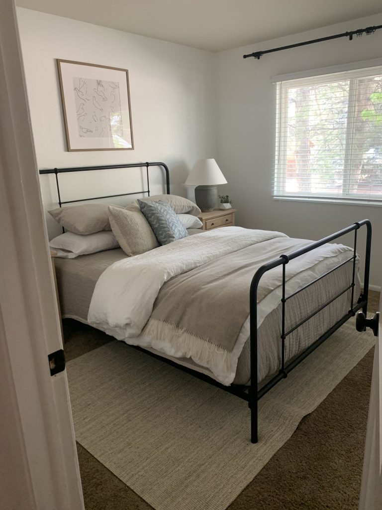 Simple minimalist bedroom with black iron bed frame, neutral layered bedding, jute area rug, light wood nightstand, and a single framed line art print on white wall