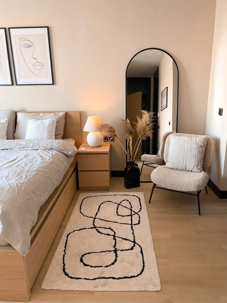 Scandi minimalist bedroom with light wood bed and nightstand, abstract rug, arched floor mirror, boucle accent chair, pampas grass in black vase, and line art wall prints