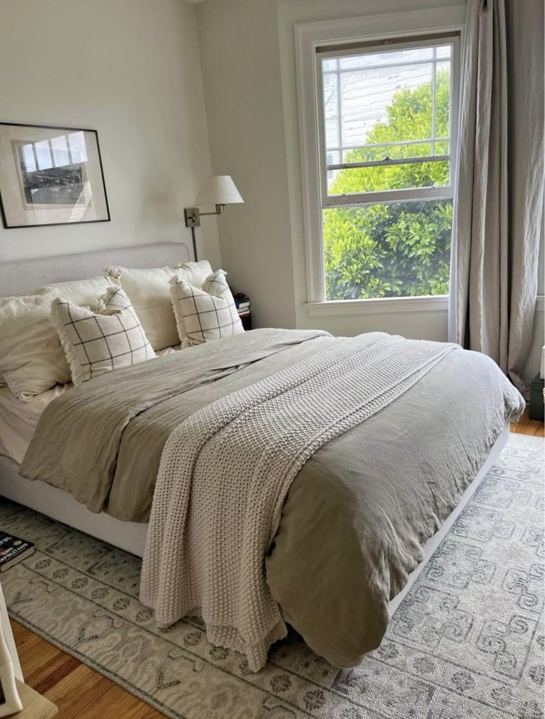 Cozy minimalist bedroom with linen duvet in warm grey, chunky knit throw, grid pattern pillow covers, wall mounted swing arm lamp, and large window with natural light
