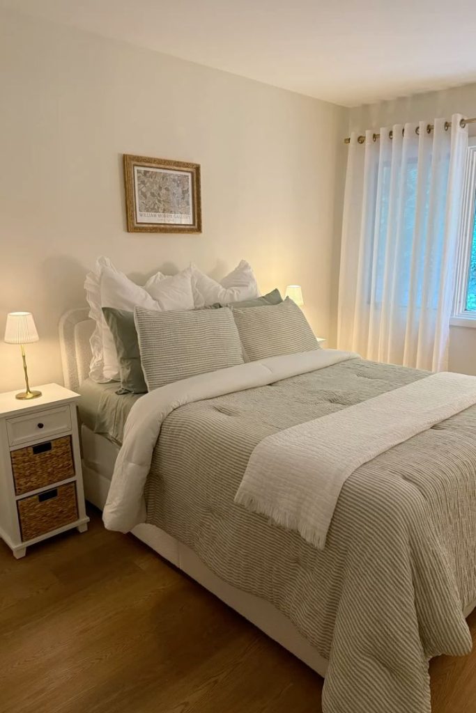 Light and airy minimalist bedroom with sage striped bedding, white upholstered headboard, white nightstand with woven basket drawers, gold framed botanical print, and sheer curtains