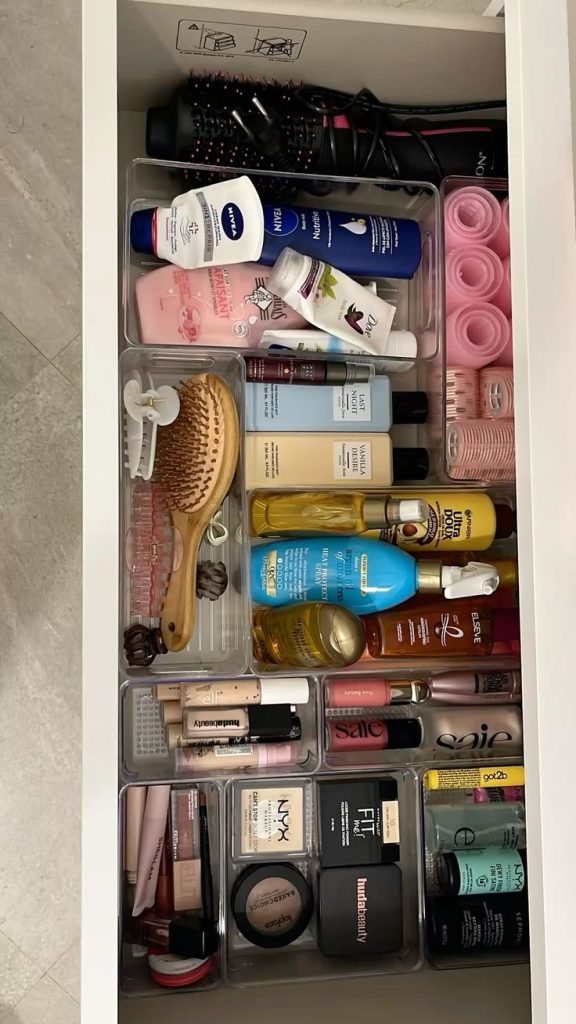 Organized bathroom drawer with clear acrylic dividers holding makeup, hair care products, brushes, and styling tools in neat sections