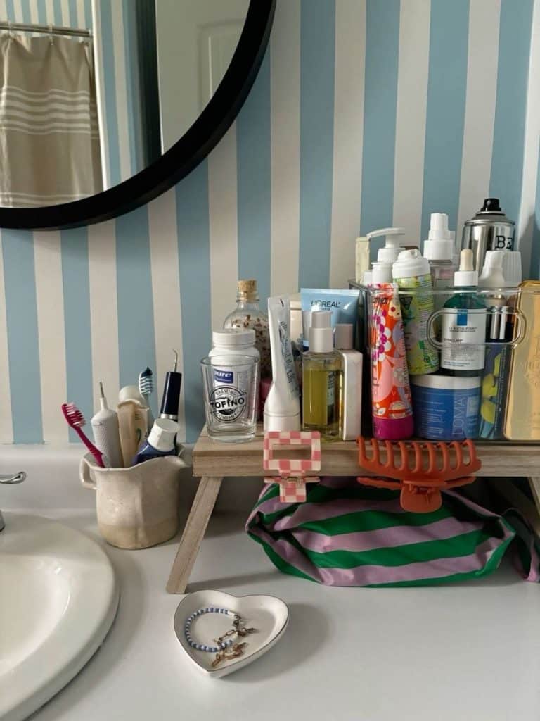 Colorful bathroom counter setup with a wooden riser shelf holding skincare and hair products, a ceramic cup with toothbrushes, and a heart-shaped jewelry dish on a blue and white striped wall background