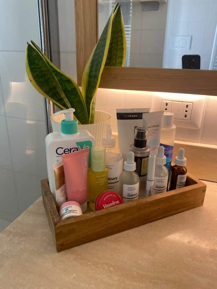 Wooden rectangular tray on a bathroom counter holding a collection of skincare products including CeraVe, The Ordinary serums, Vaseline, and other face care items alongside a potted snake plant