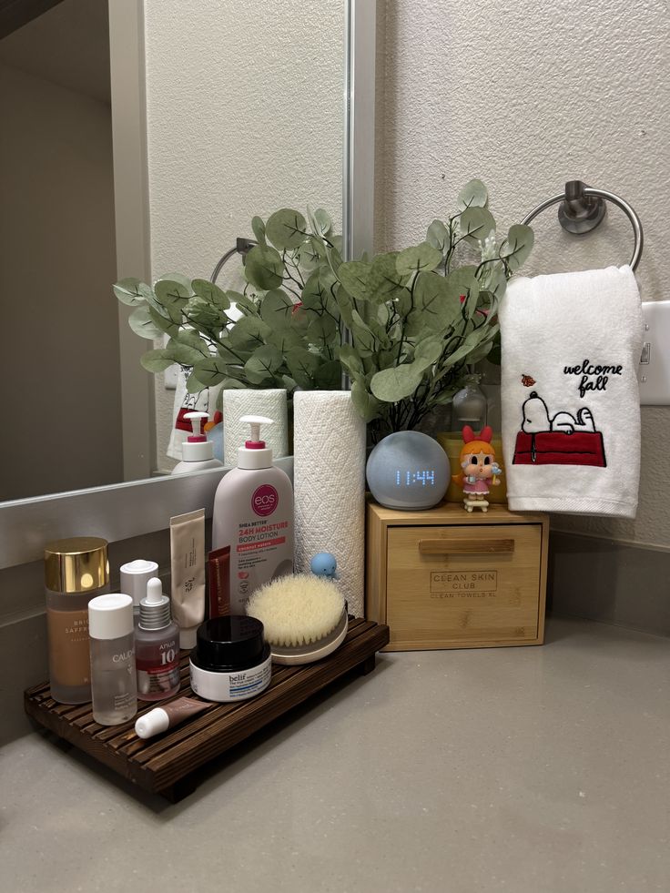 Bathroom counter corner with a dark wood riser tray holding skincare products and a facial brush, next to a bamboo drawer unit and a eucalyptus branch arrangement in a white vase