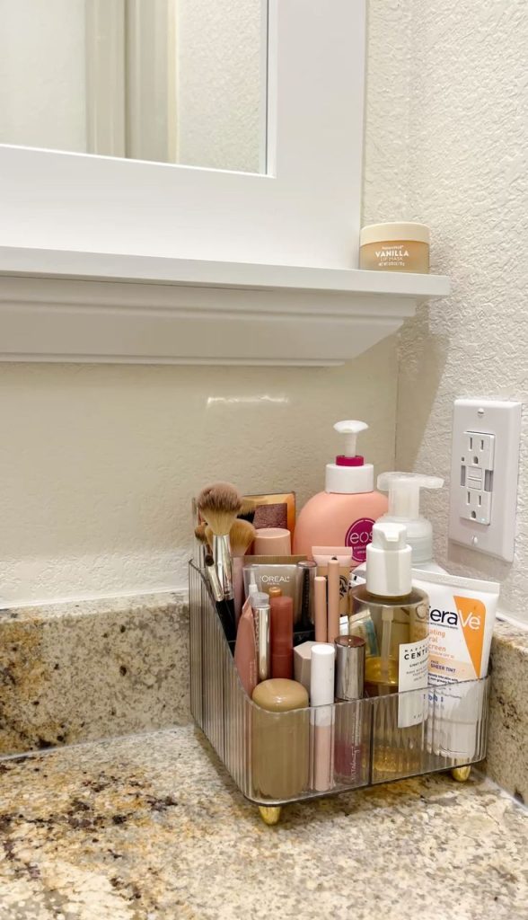 Clear ribbed acrylic makeup and skincare caddy with gold feet on a granite bathroom counter holding brushes, lipstick, foundation, and skincare products in divided compartments
