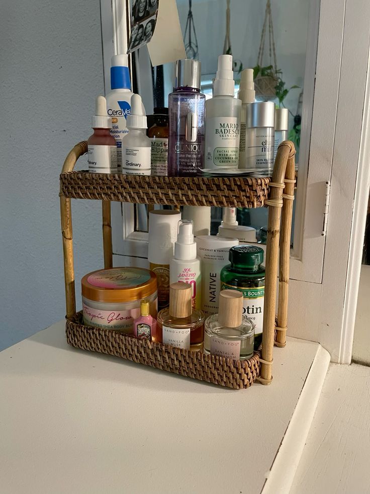 Rattan two-tier bathroom countertop shelf holding skincare bottles, Clinique makeup remover, Mario Badescu spray, CeraVe, body butter, perfume, and deodorant on both levels