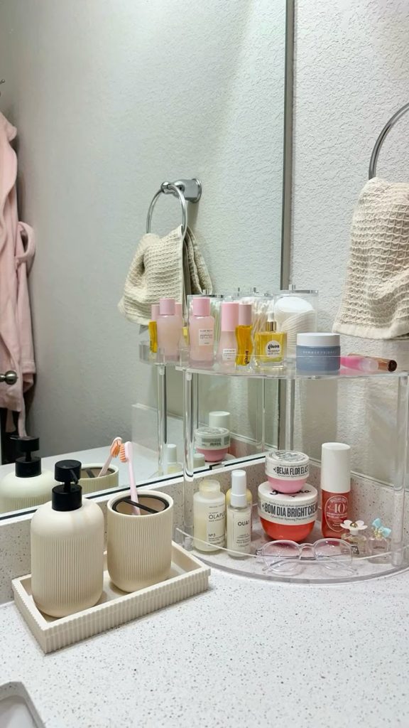 Clear acrylic two-level rotating lazy Susan bathroom organizer holding Gisou hair oil, Glow Recipe toner, Sol de Janeiro products, and skincare creams, alongside a cream ribbed tray with soap dispenser and toothbrush cup