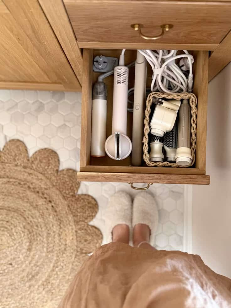 Warm oak bathroom drawer with a hair dryer, curling wand, and accessories stored neatly alongside a small woven basket holding attachments