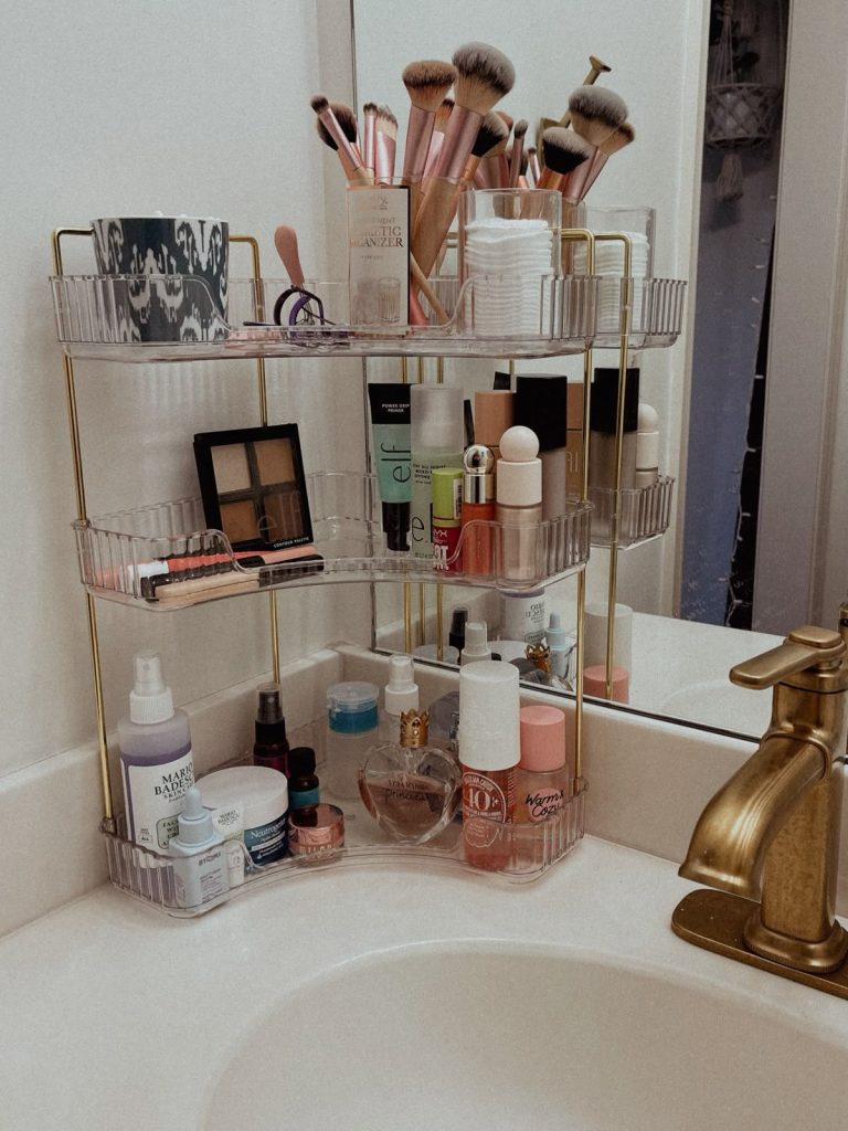 Three-tier clear acrylic and gold corner shelf organizer on a bathroom vanity holding makeup brushes in cups, eyeshadow palettes, skincare products, perfume, and nail polish