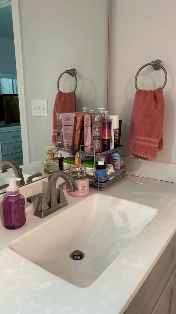 Two-tier metal wire bathroom countertop organizer holding Bath and Body Works body care products, perfume bottles, and skincare items next to a sink with mauve towels in the background