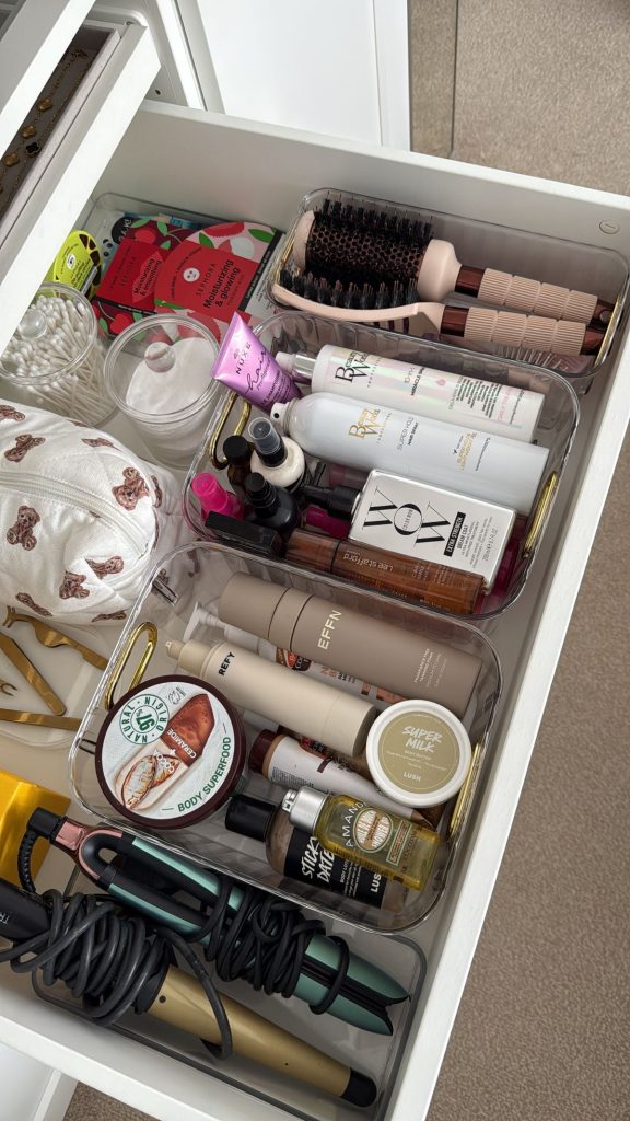 White vanity drawer organized with clear acrylic bins and cotton rope baskets holding hair brushes, hair products, body care, and styling tools