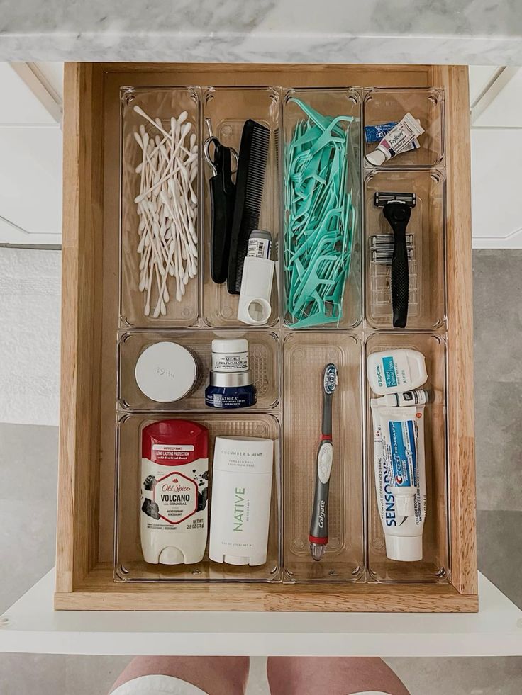 Bathroom drawer with clear acrylic organizers holding cotton swabs, dental floss, deodorant, toothbrushes, razors, and other daily essentials in separate zones