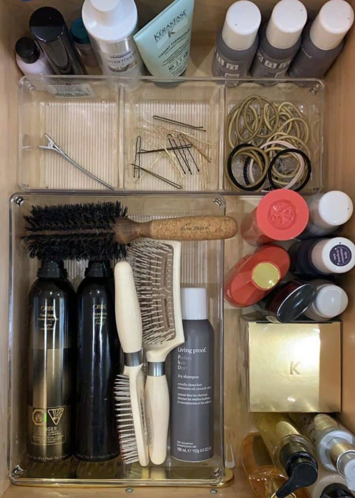 Bathroom drawer with clear acrylic bins holding boar bristle brushes, dry shampoo, hair styling products, bobby pins, and hair ties in organized sections