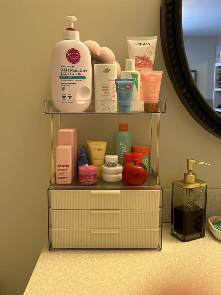 Clear acrylic two-tier bathroom countertop shelf with gold accents holding skincare and body care products on the upper level and face creams and lip masks on the lower level, with small pull-out drawers at the base